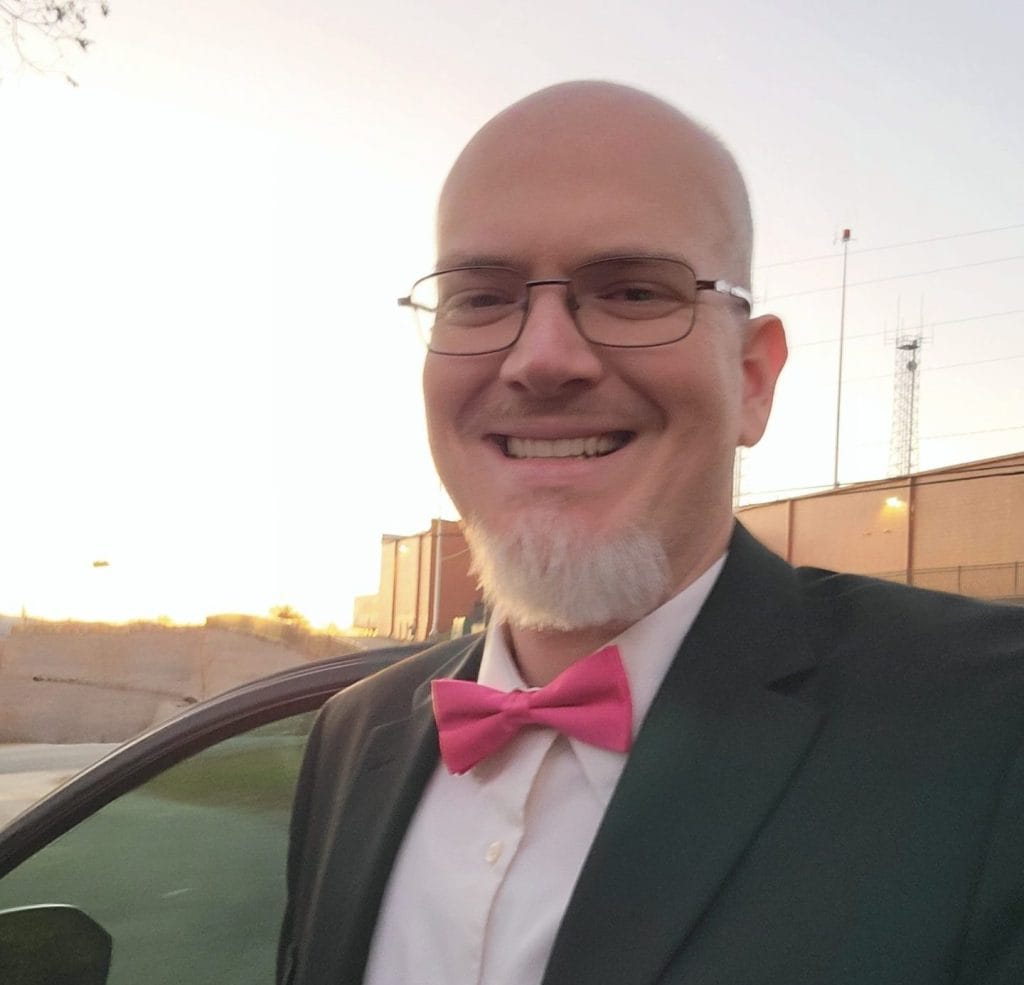 Smiling man in formal attire with a pink bowtie outdoors at sunset.